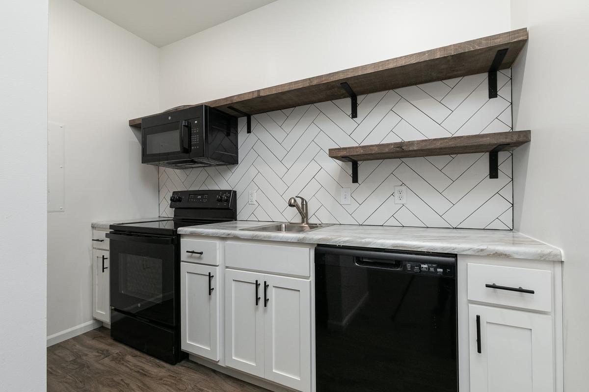 Vacant kitchen: white cabinets, black range, microwave and dishwasher, open shelving, herringbone tile