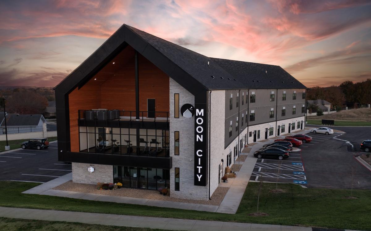 Aerial of the building at dusk from the front: gabled glass-fronted entrance, MOON CITY sign, parking lot, pink sky