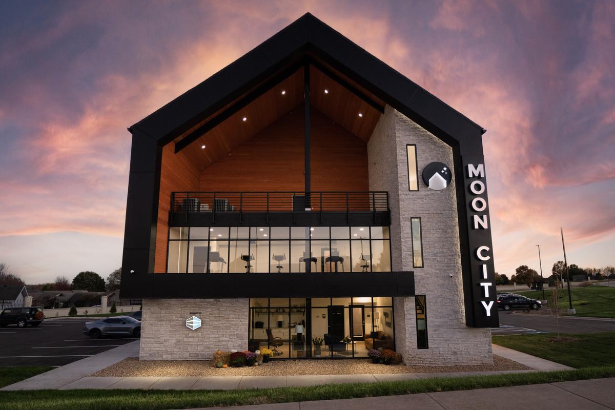 Front elevation at dusk: gable, glass lobby, wood soffit, MOON CITY sign, lit windows